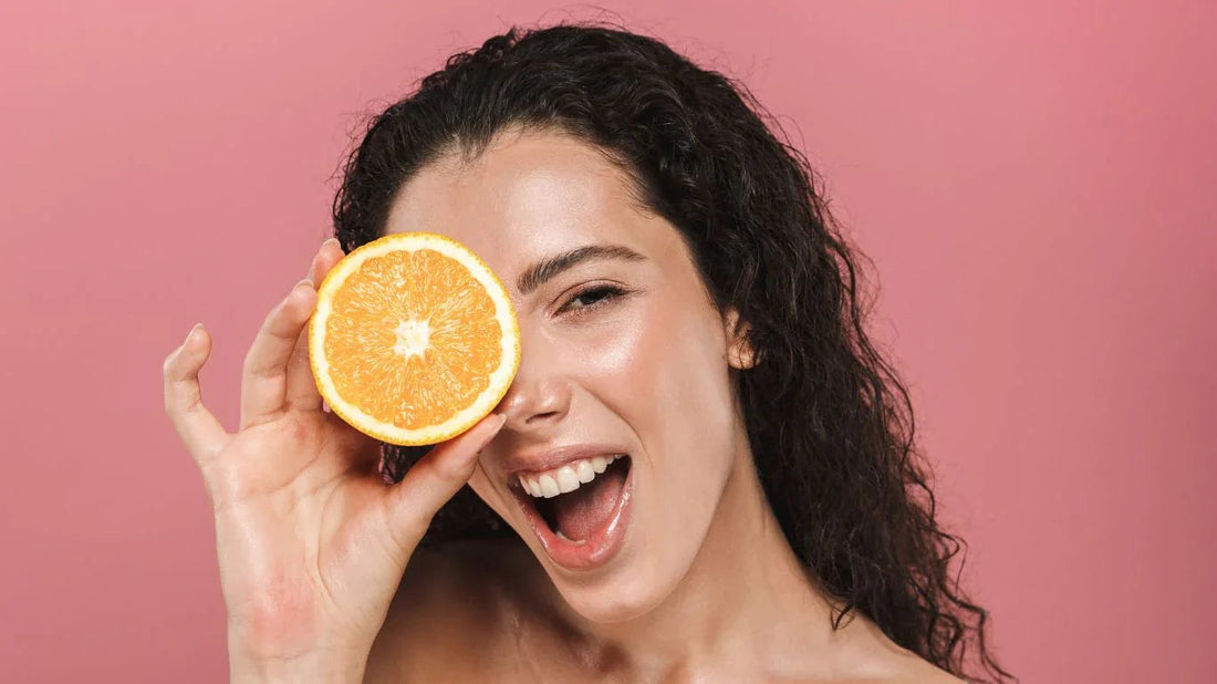 Smiling woman with curly hair holding half of a fresh orange against her eye over pink background