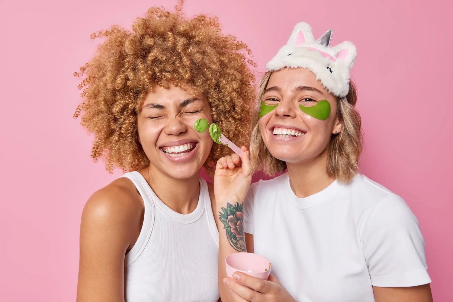 Two smiling women applying green face masks, one with curly hair in white tank top and another with short hair wearing unicorn eye mask