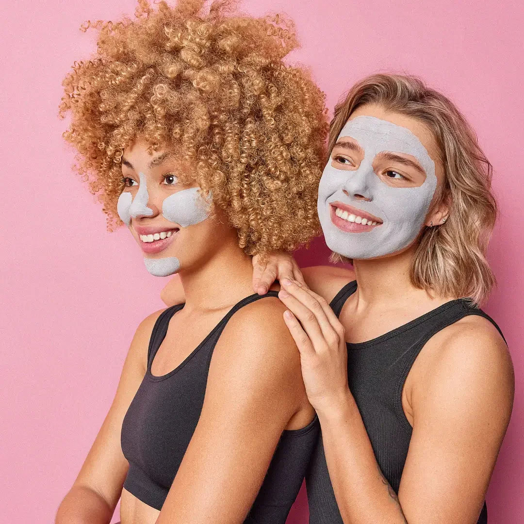 Two smiling women in black tank tops with gray face masks against pink background, showing skincare routine